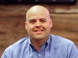 Headshot of Jerry Beighey wearing a button-down shirt, smiling in front of a blurred wood background.