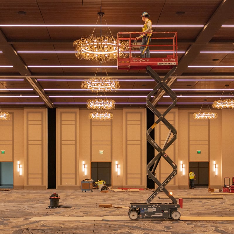 A worker on a red scissor lift adjusts a chandelier in a large ballroom, with other workers and construction materials on the floor