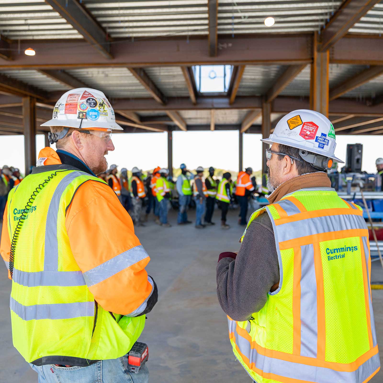 Two construction workers in hard hats and safety vests converse on a building site with a group of workers in the background