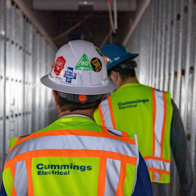 Two construction workers in safety vests walking down a hallway, discussing their tasks as they proceed