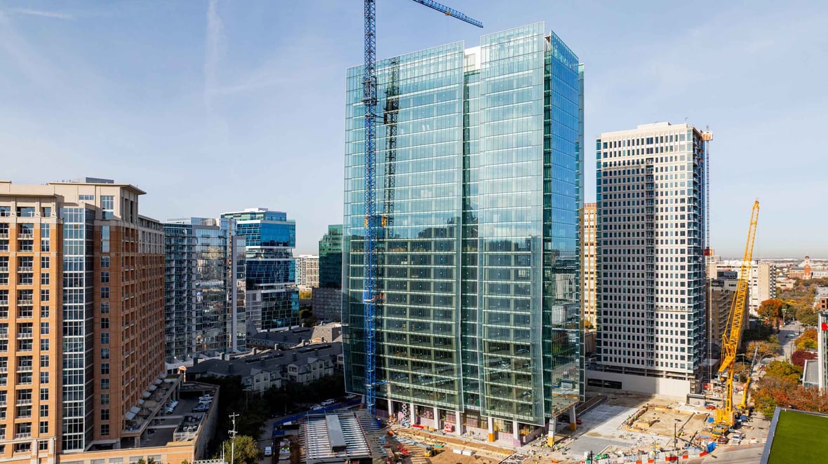 Glass office tower under construction in a dense downtown setting with cranes and surrounding high-rises.
