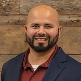 Headshot of Abraham Gonzalez wearing a button-down shirt and black blazer, smiling in front of a wood background.