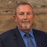 Headshot of Ben Michener wearing a button-down shirt with a tie and black blazer, smiling in front of a wood background.