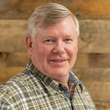 Headshot of Chris Carroll wearing a button-down shirt, smiling in front of a wood background.