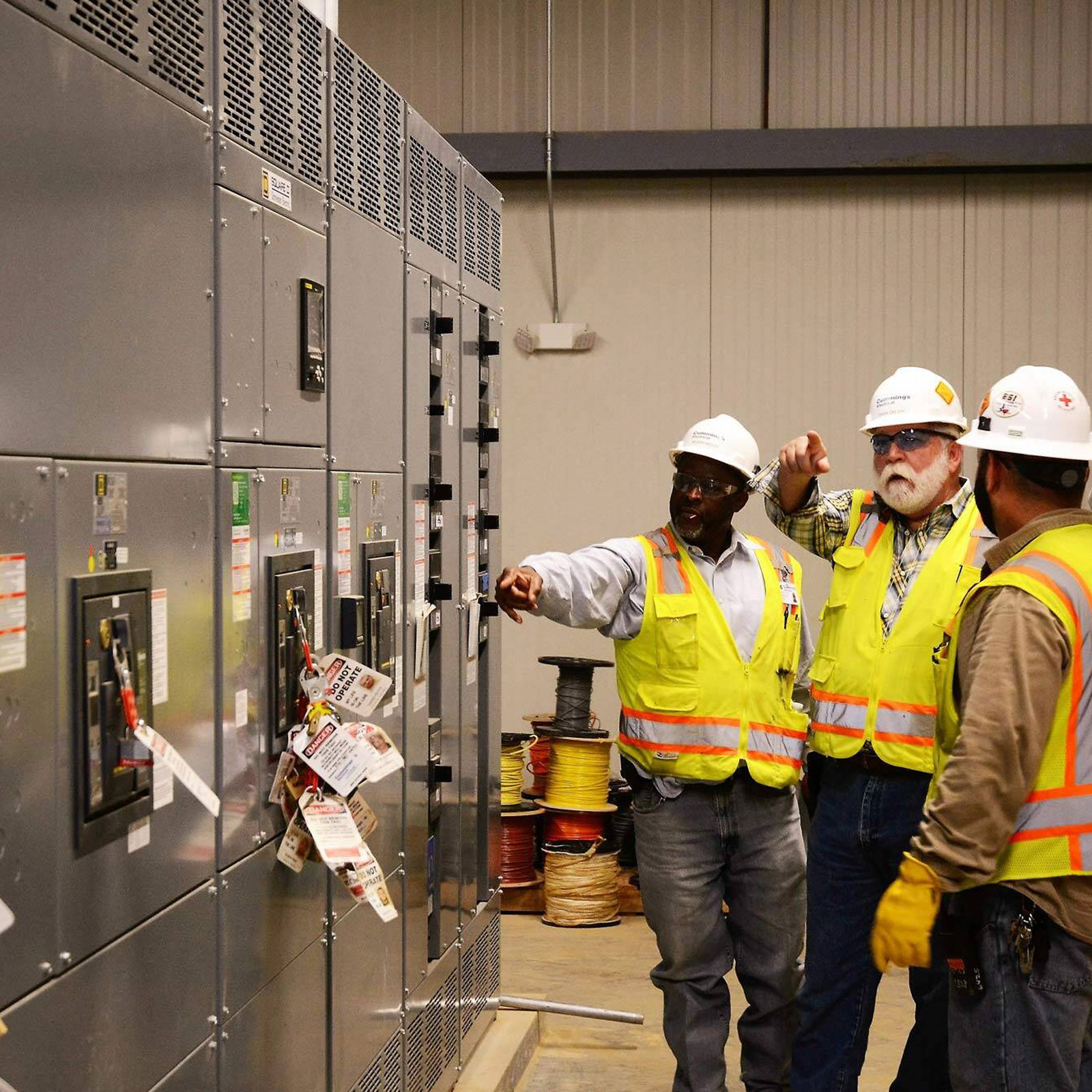 3 men with PPE evaluating electrical panels pointing in various directions
