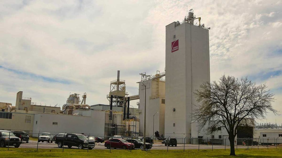 Ground-level view of the Owens Corning manufacturing plant with tall processing towers, support buildings, and a fenced perimeter with parking