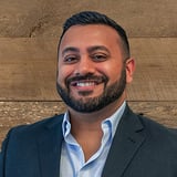 Headshot of Humza Khan wearing a button-down shirt and black blazer, smiling in front of a wood background.