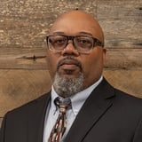 Headshot of Jason Puryear wearing a button-down shirt with a tie and black blazer, in front of a wood background.
