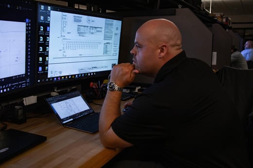 View of a man analyzing electrical drawings on a large monitor at a desk with a laptop open in front of him.