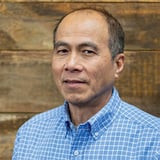 Headshot of Phu Tran wearing a button-down shirt, smiling in front of a wood background.