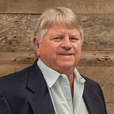 Headshot of Steve Lemay wearing a button-down shirt and black blazer, smiling in front of a wood background.