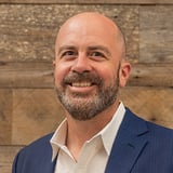 Headshot of Will Keeney wearing a button-down shirt and blue blazer, smiling in front of a wood background.